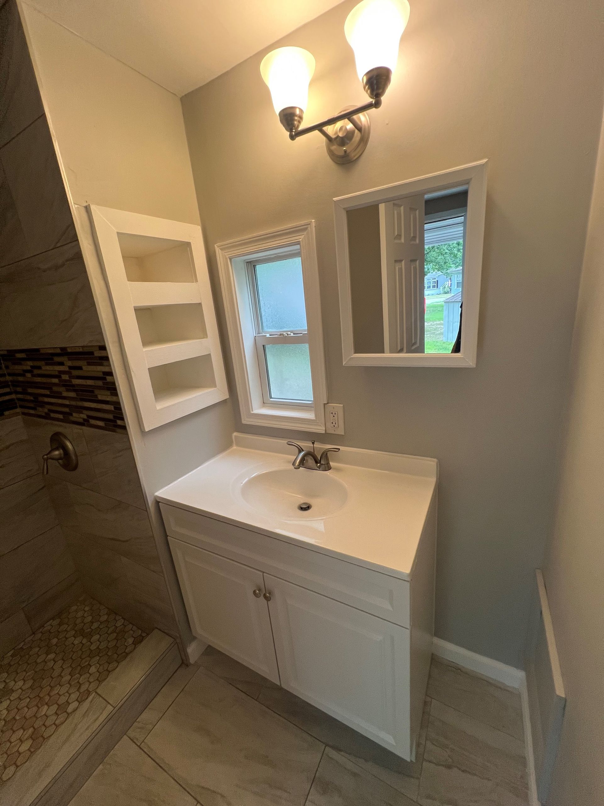 Bathroom vanity with white sink, mirror, wall sconce, and open window beside tiled shower