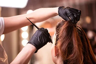Gloved hands applying hair dye to a client’s brown hair in a salon