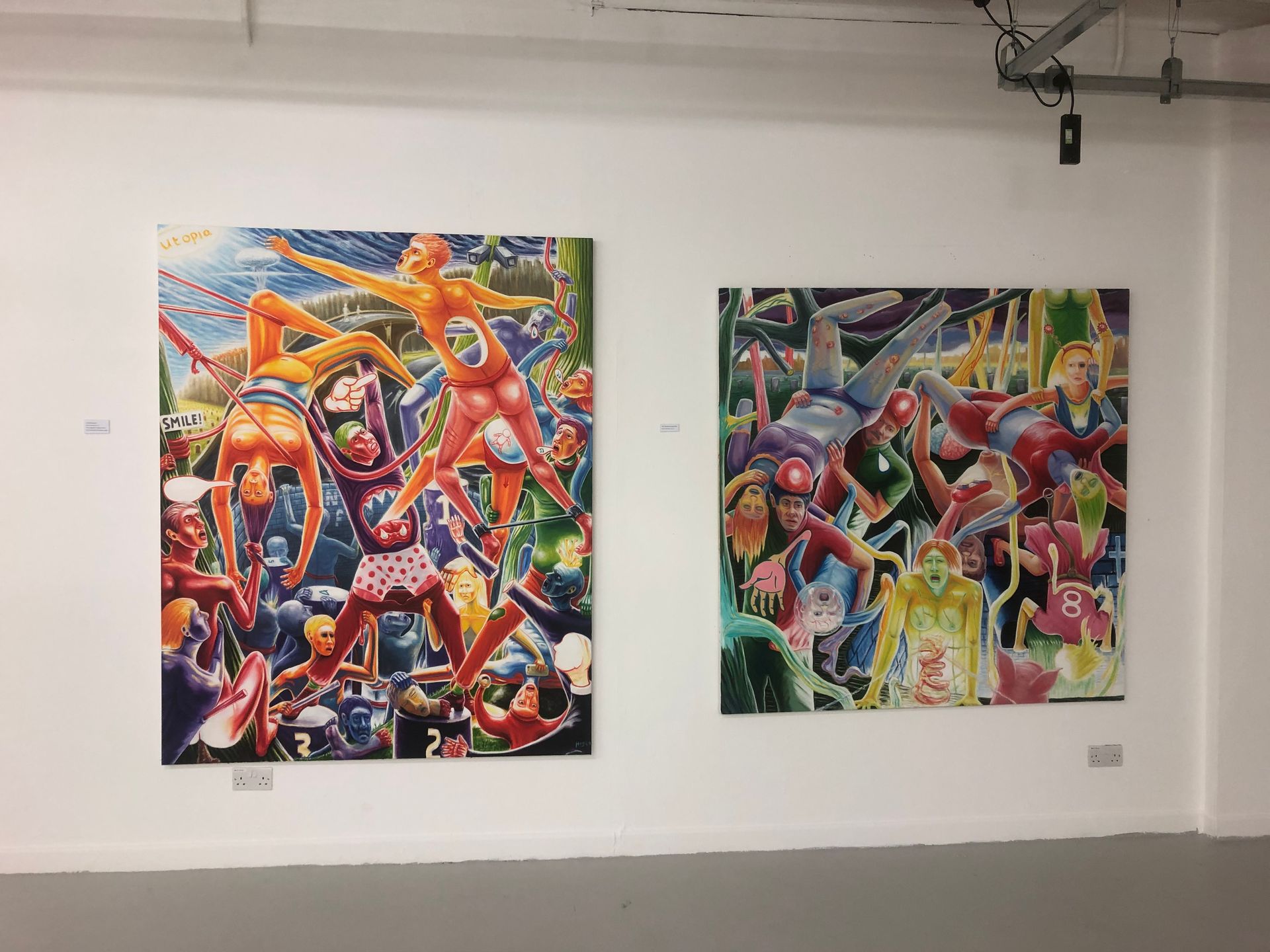 Two colorful, surreal paintings hanging on a white wall in an art gallery.
