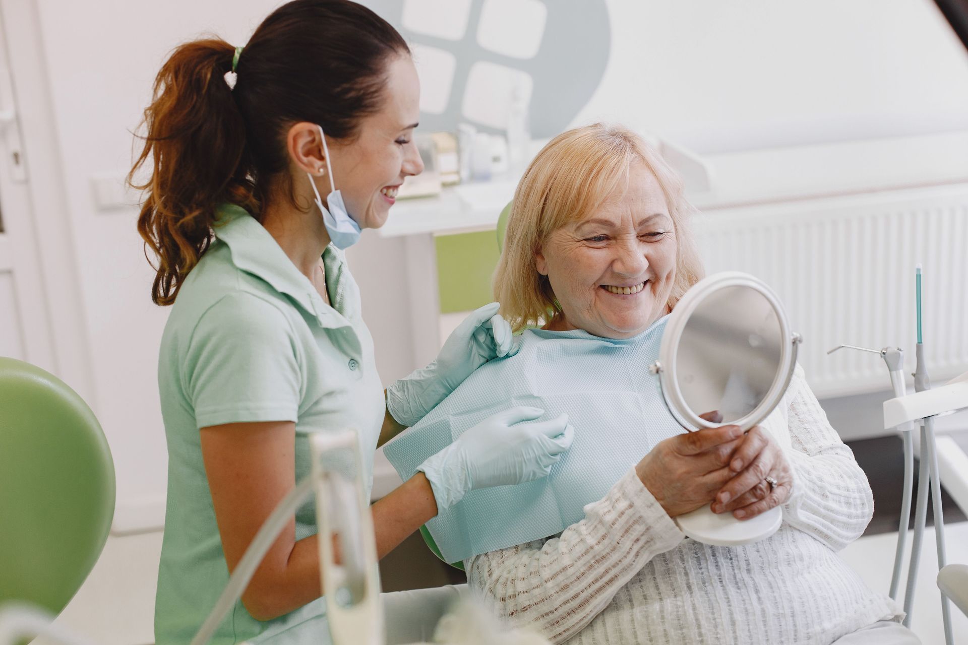 Patient sitting down in dental chair holding a mirror with the dentist next to them checking results