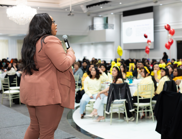 Anisha Angella standing on stage performing a workshop in front of a large crowd seated at dining tables