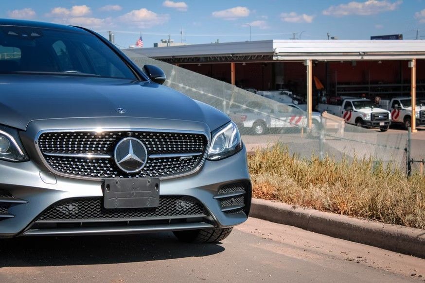 A silver Mercedes-Benz parked on the side of a road with a building and service trucks in the background.