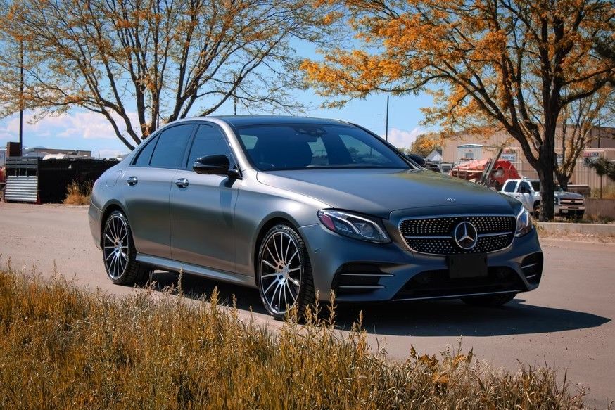 A grey Mercedes-Benz sedan parked on a dirt road, surrounded by trees with autumn foliage under a clear blue sky.