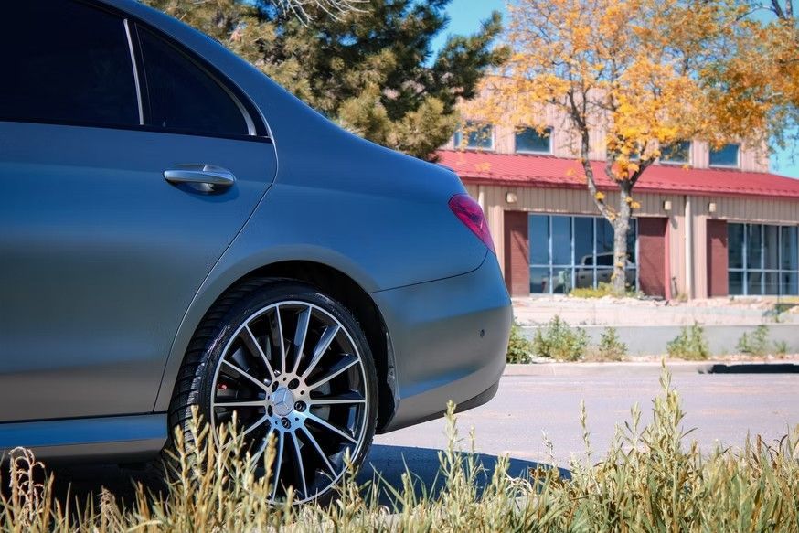 A side view of a matte gray luxury sedan parked in front of a building with a red roof, framed by autumn trees and grass.