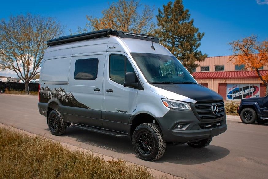 Silver Mercedes-Benz camper van with custom mountain decals, lifted suspension, and black trim parked on an asphalt lot.