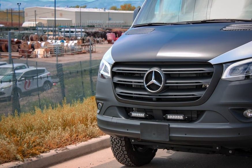 A close-up view of the front of a matte grey Mercedes-Benz Sprinter van parked on an asphalt road.