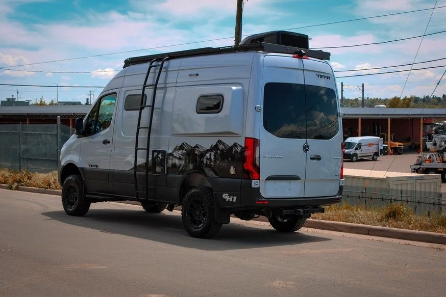 A silver camper van with a mountain-themed decal and roof ladder parked on an asphalt street.