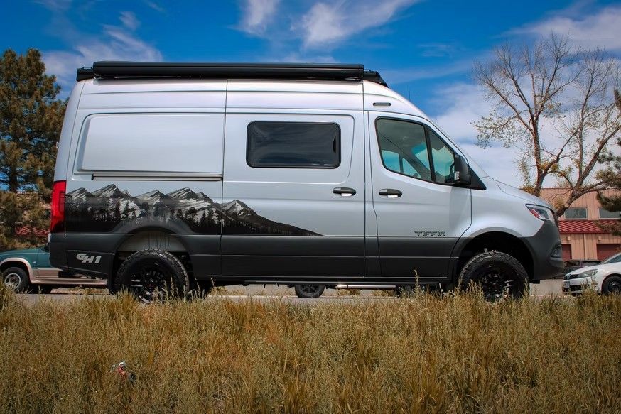 A silver adventure van with a black mountain range decal parked in a field under a blue sky.