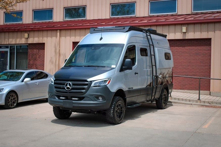 A silver and black Mercedes-Benz Sprinter camper van with off-road tires and a roof ladder parked in a paved lot.
