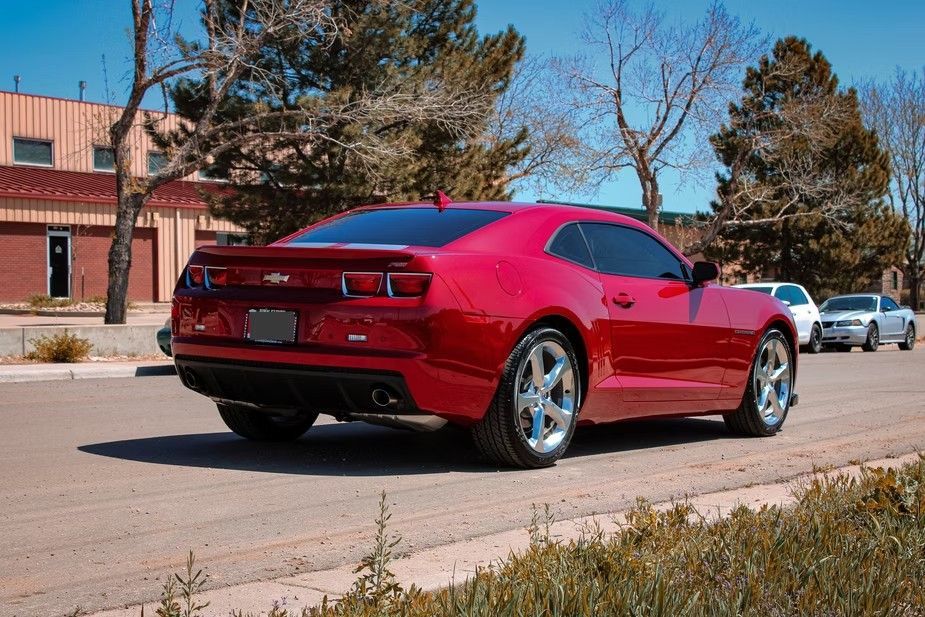 A red Chevrolet Camaro parked on a street near a building with trees in the background on a sunny day.