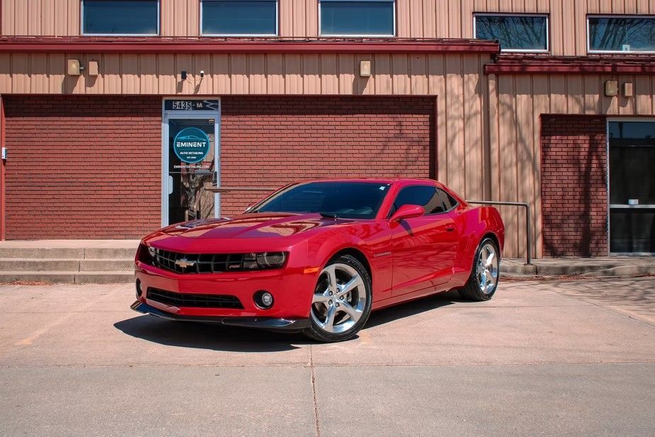 A bright red Chevrolet Camaro parked on a concrete lot in front of a brick building with windows.