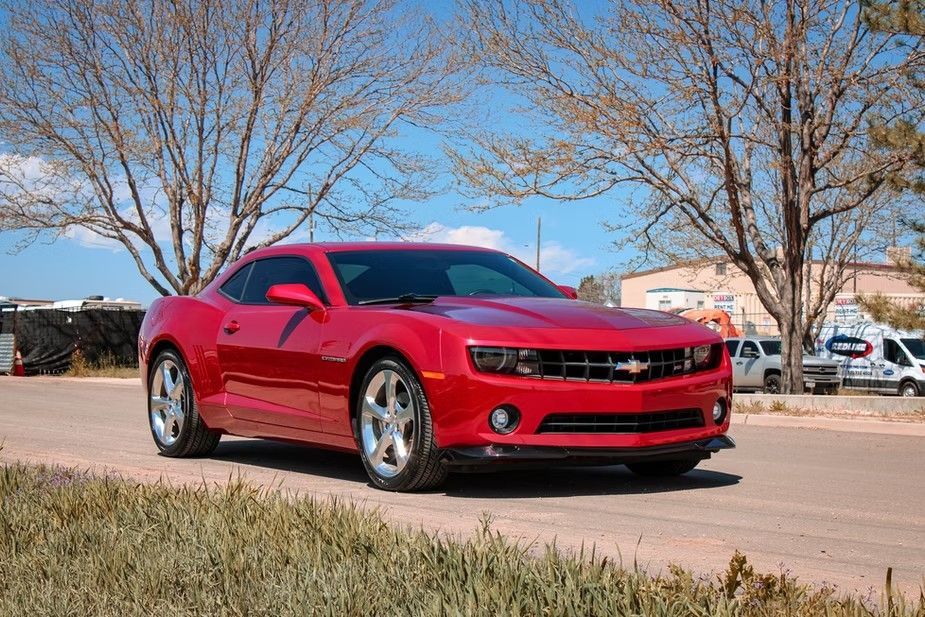 A bright red Chevrolet Camaro parked on an asphalt road in front of bare trees under a clear blue sky.
