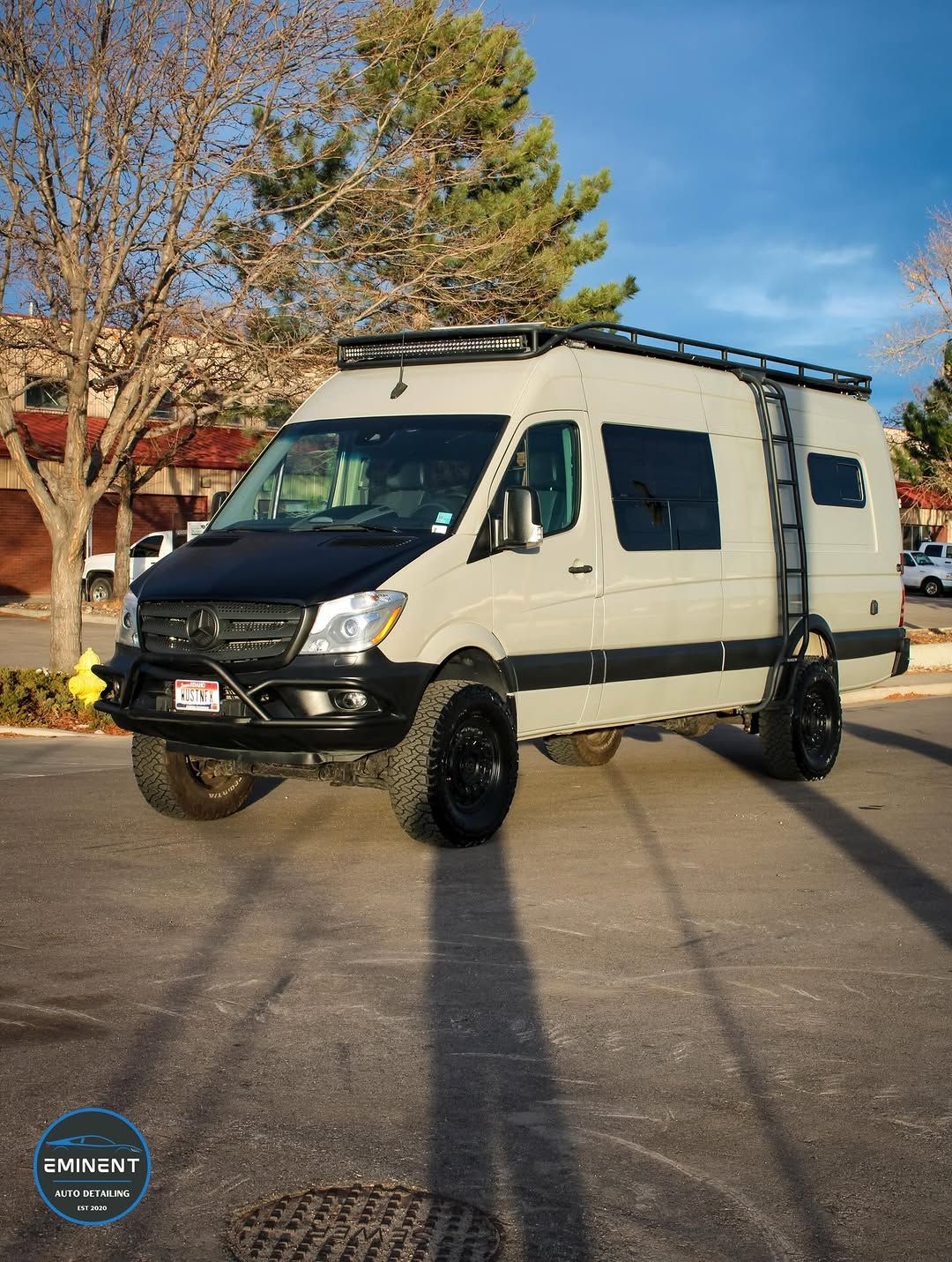 Off-road van with roof rack, ladder, and black accents parked on asphalt.