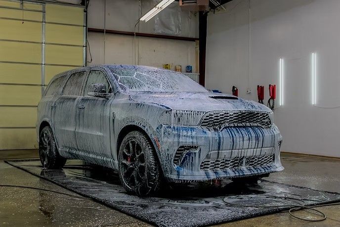 A dark-colored SUV covered in thick white cleaning foam, parked inside a garage wash bay.