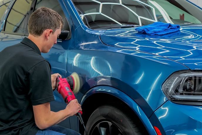 A person uses a red power buffer to polish the blue front fender of a vehicle in a well-lit workshop.