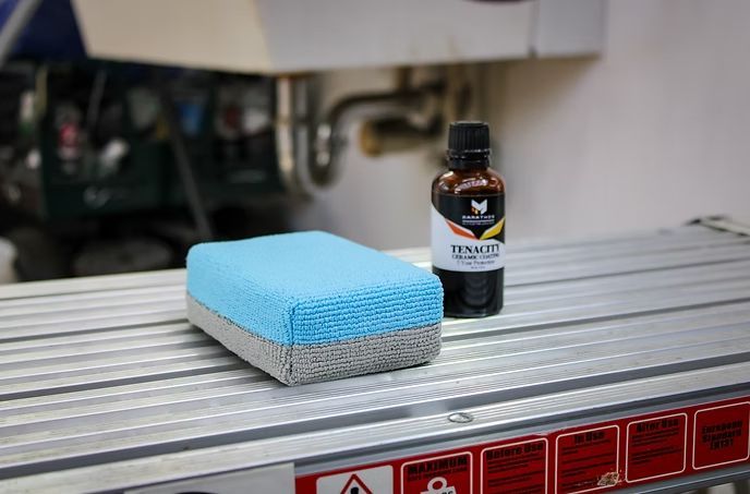A blue and grey sponge sits next to a small bottle of Renacit cleaner on a metal surface.