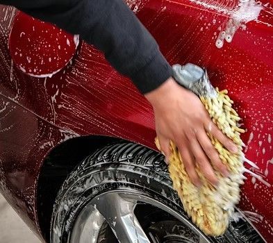 A hand uses a yellow microfiber wash mitt to clean the red side panel of a car above a tire.