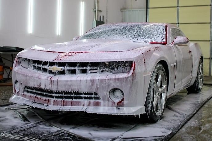 A Chevrolet Camaro covered in white soap foam during a professional car wash inside a garage.