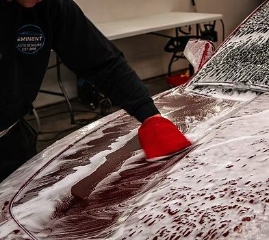 A person wearing a black sweatshirt washes a red car with a red microfiber mitt in a garage.