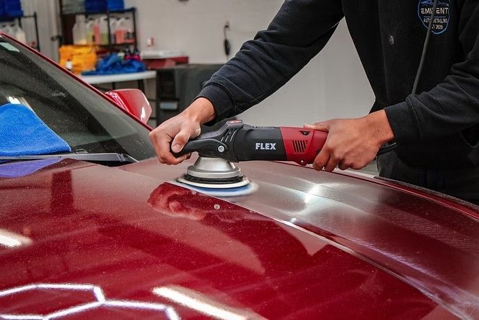 A person uses a red Flex dual-action power polisher to buff the hood of a shiny, maroon car in a workshop.