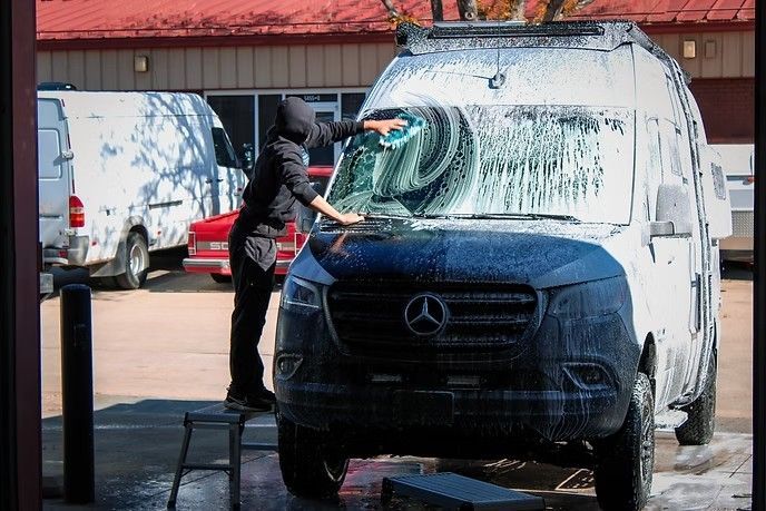 A person on a small step stool uses a squeegee to wash soap off the windshield of a black Mercedes cargo van.