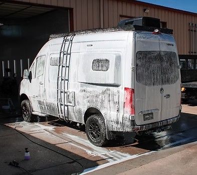 A white camper van covered in soapy foam is parked on a concrete lot during a wash.