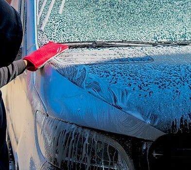 A hand wearing a red wash mitt scrubs soap suds off the hood of a blue car.