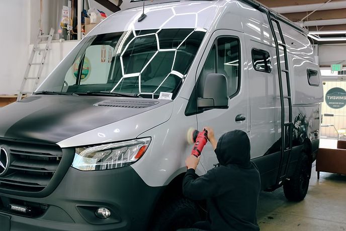 A person in a black hoodie uses a power buffer to polish the side of a silver Mercedes-Benz Sprinter van in a workshop.
