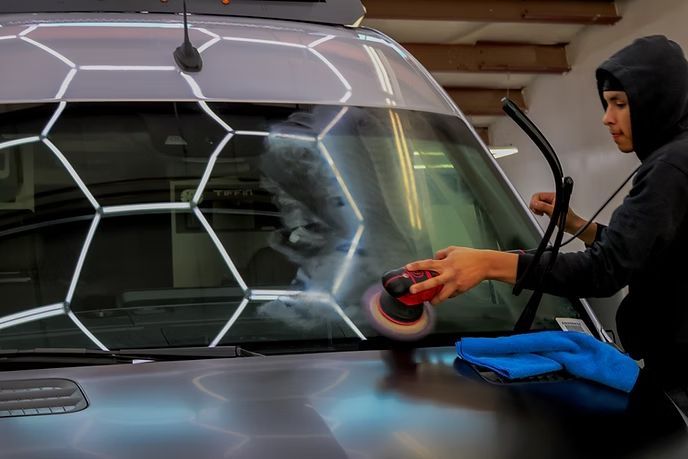 A person in a hoodie uses a power polisher to clean the windshield of a dark vehicle under hexagon-patterned ceiling lights.
