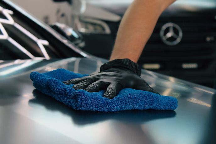 A gloved hand uses a blue microfiber towel to buff the hood of a car.