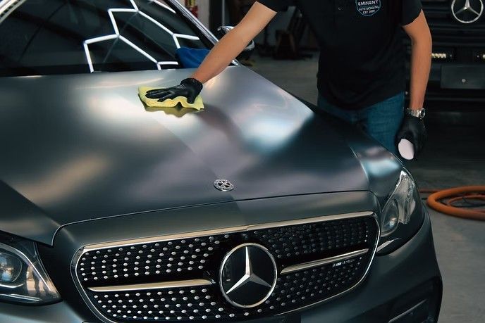 A person wearing black gloves wipes the matte dark grey hood of a Mercedes-Benz with a yellow cloth in a workshop.