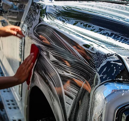 A person washes the black fender of a vehicle with a red cloth and soapy water.