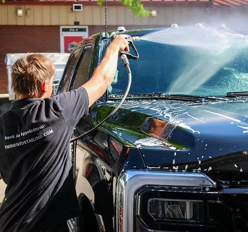 A person in a black work shirt uses a pressurized water sprayer to clean the hood and windshield of a black pickup truck.