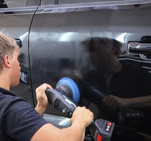 A person using an electric orbital polisher to buff the dark, glossy door panel of a vehicle.
