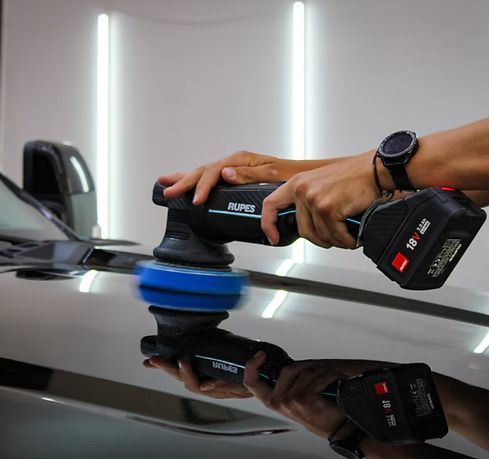 A person uses a handheld cordless RUPES polisher to buff a shiny black car hood in a brightly lit garage.