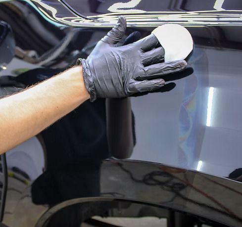 A gloved hand uses a white applicator pad to apply protective coating to a shiny black car panel in a workshop.
