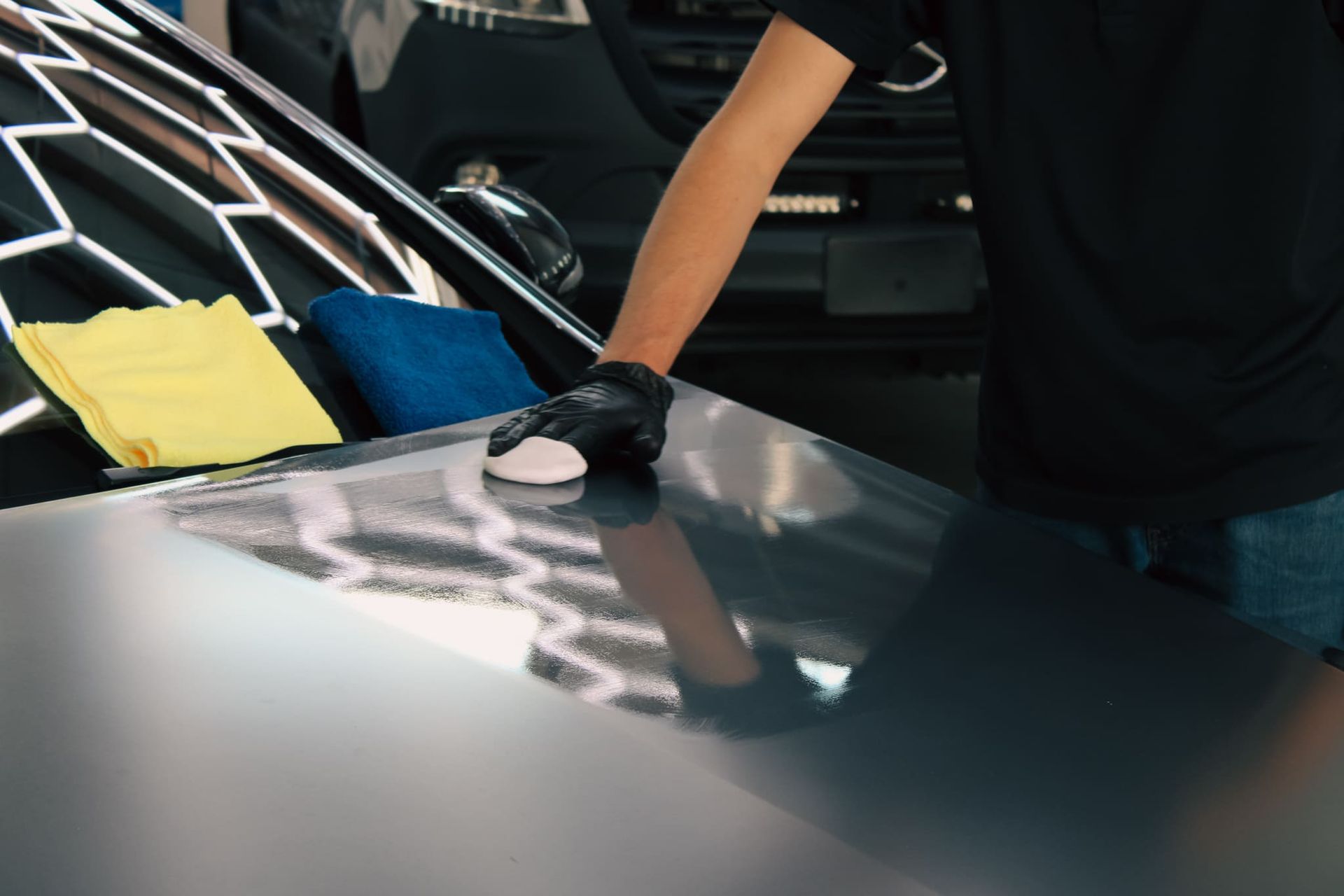 Person in black glove waxing a car hood with a white applicator pad, blue and yellow cloths nearby.
