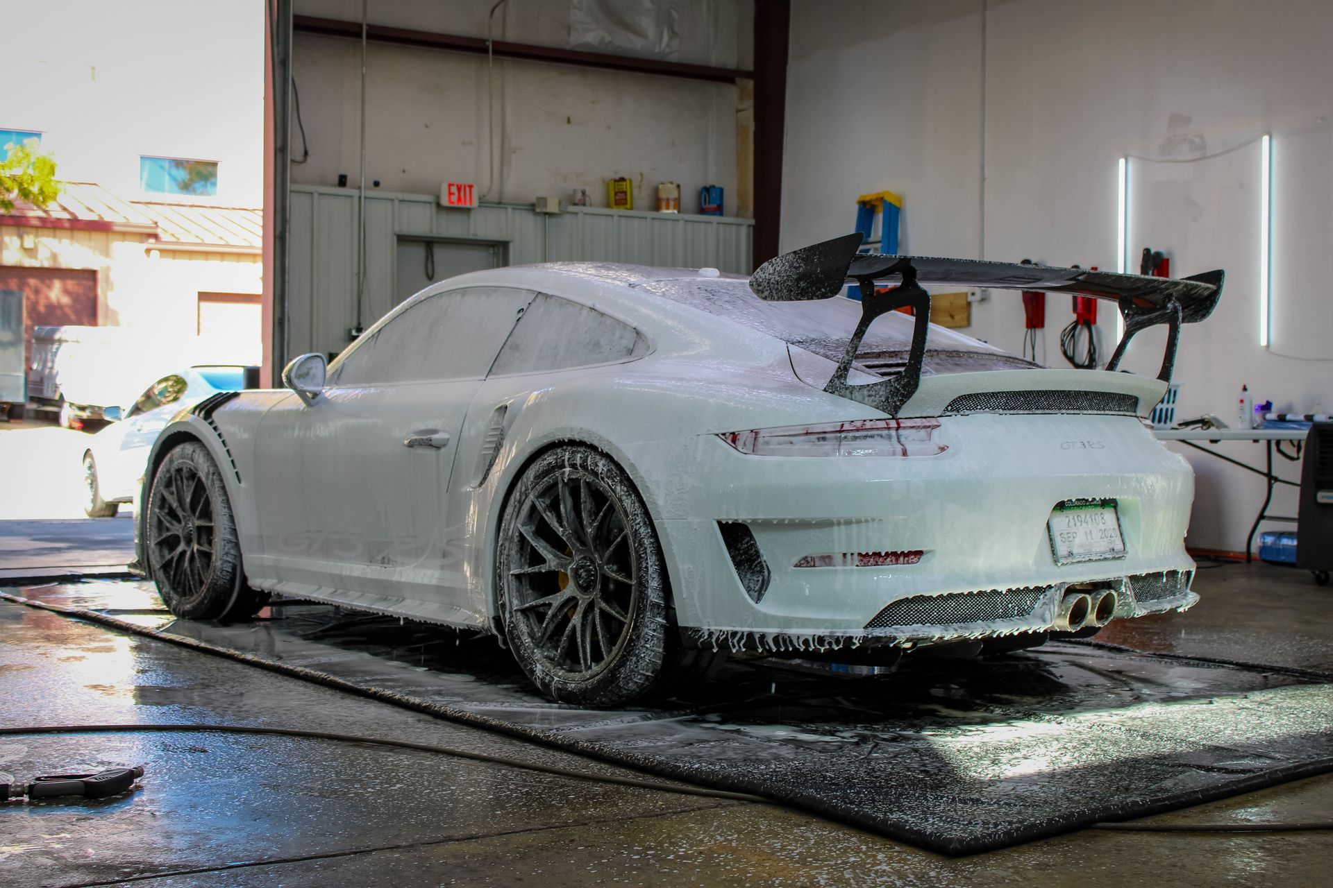 White Porsche sports car covered in foam suds, being washed in a garage with black tires and a large rear wing.