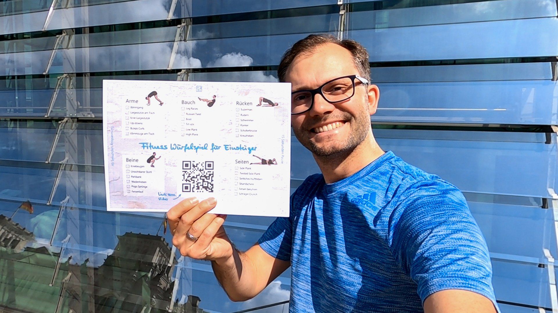 Ein Mann mit blauem T-Shirt hält das Poster vom Fitness Würfelspiel