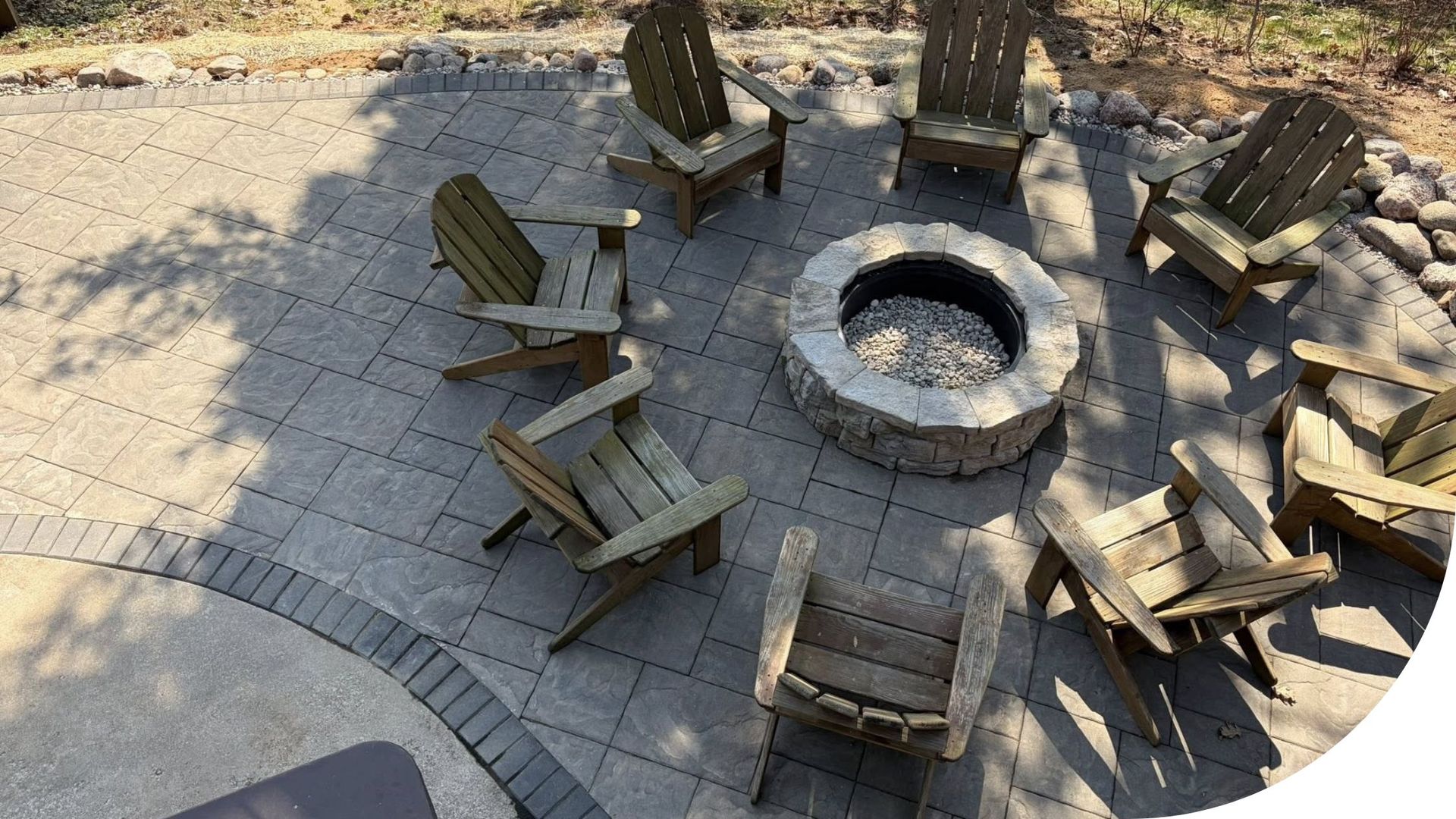 Outdoor stone fire pit surrounded by wooden chairs on a paved patio in sunlight
