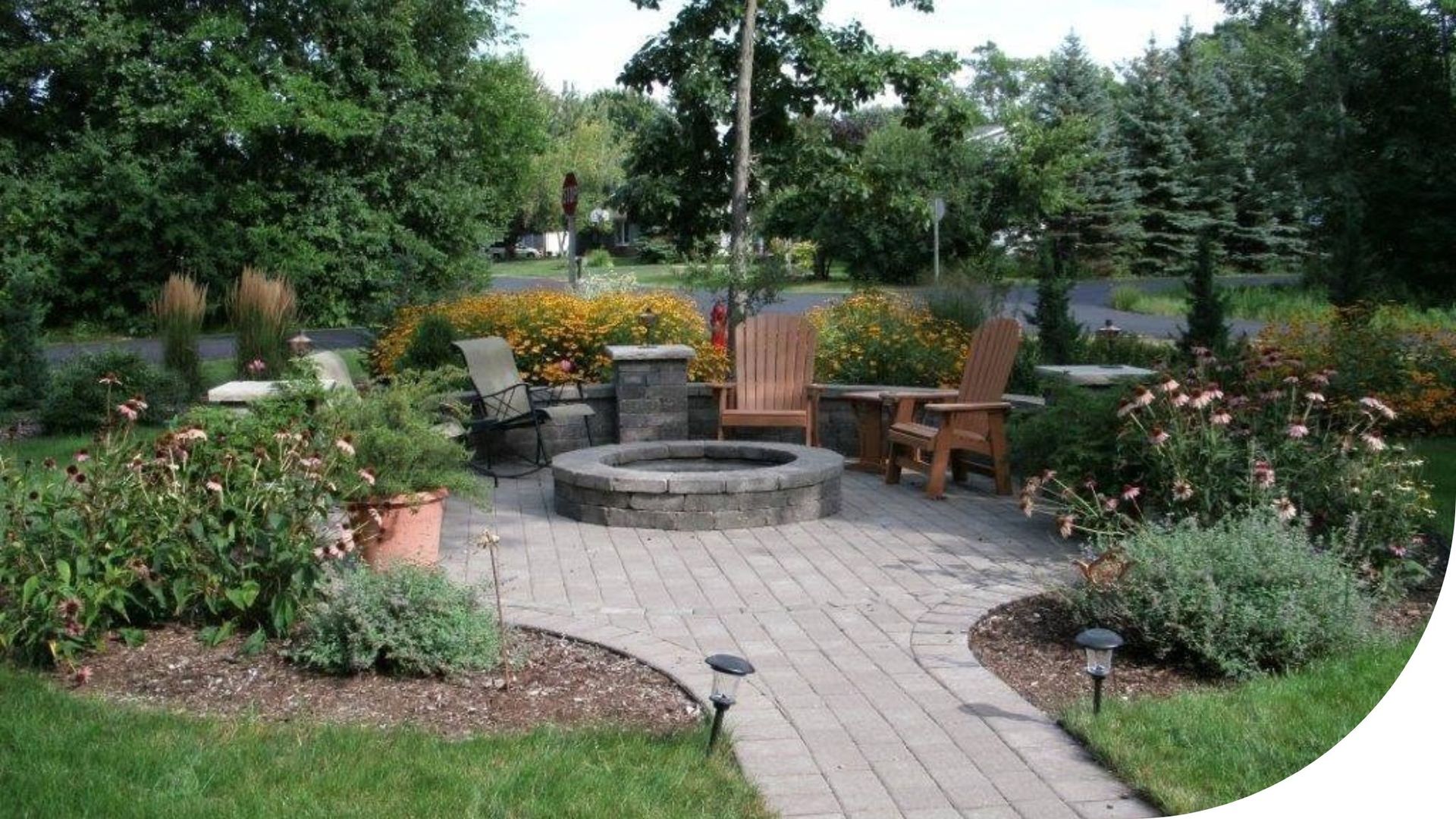 Garden patio with stone fire pit, wooden chairs, and surrounding flowers and shrubs