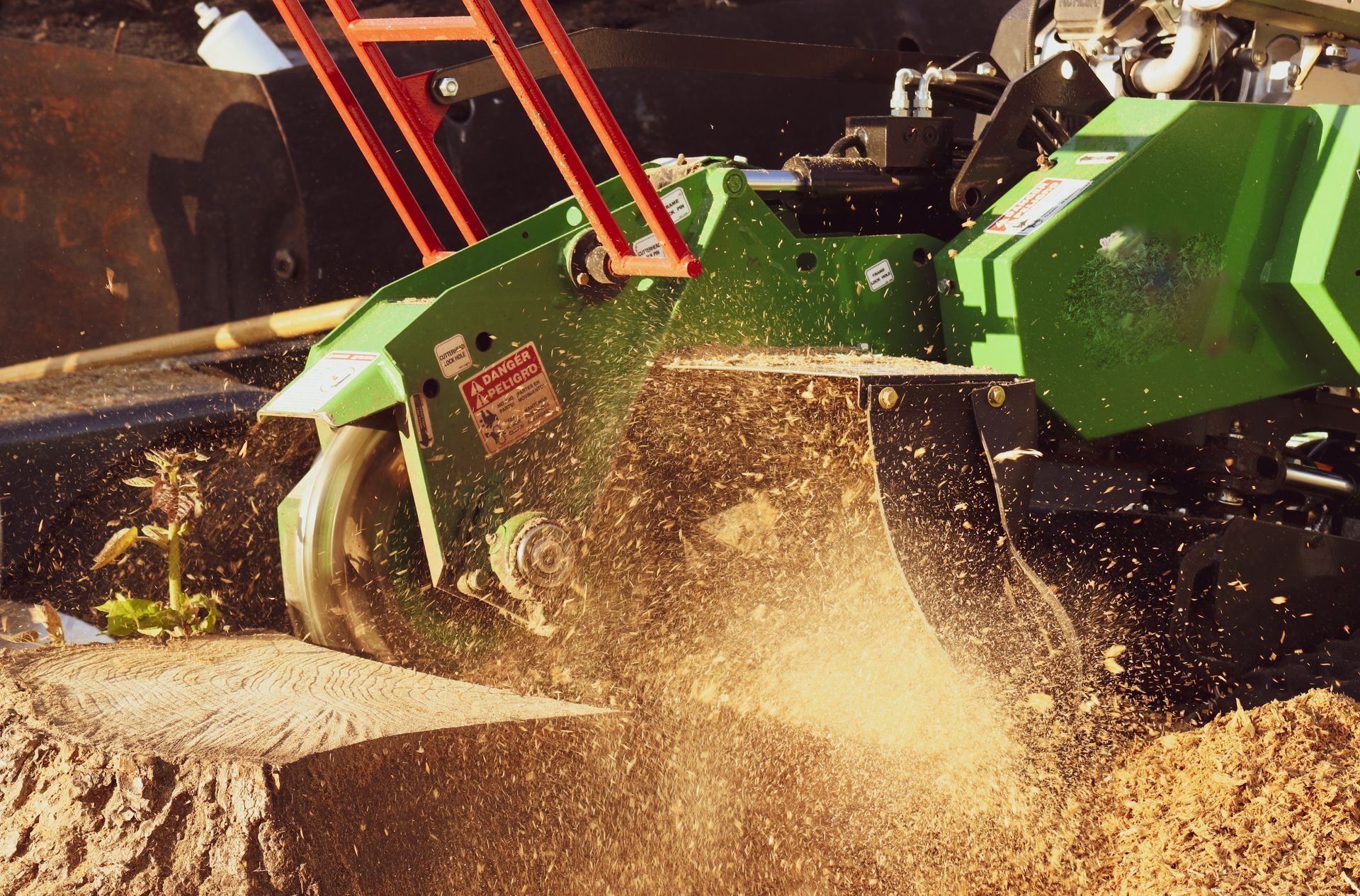 A bright green stump grinder cuts into a tree stump, creating a cloud of wood chips.