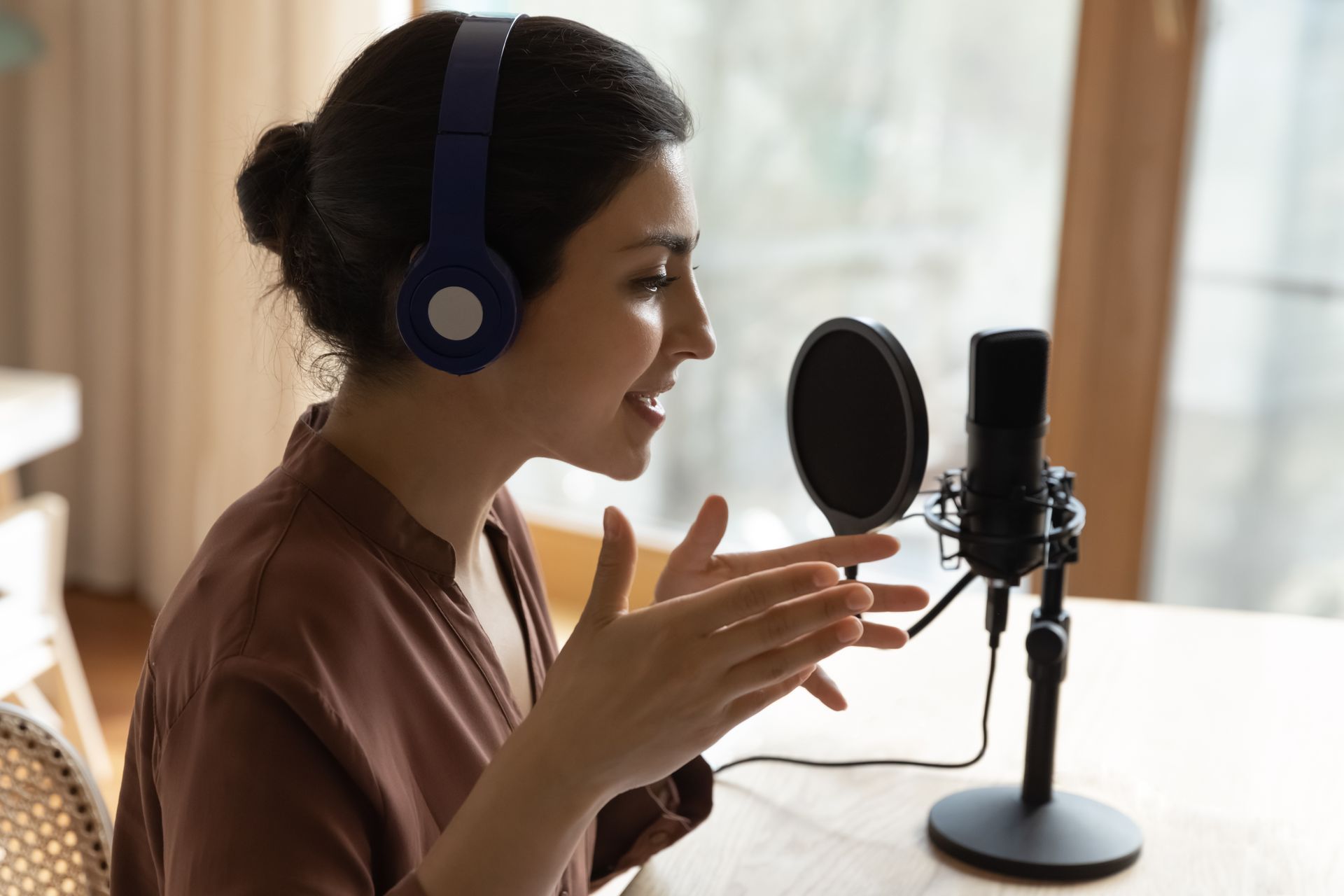 A person wearing blue headphones talks into a microphone with a pop filter while recording in a bright, indoor setting.