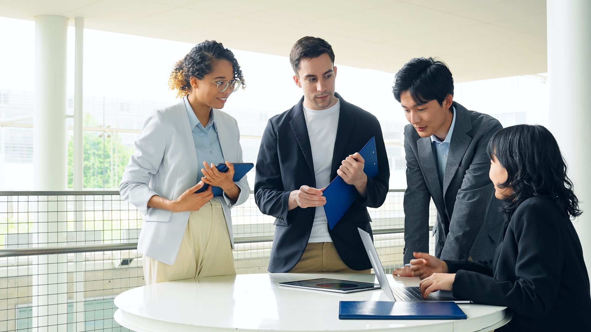 Four professionals collaborate around a circular table with a laptop, documents, and folders in a modern, bright office.