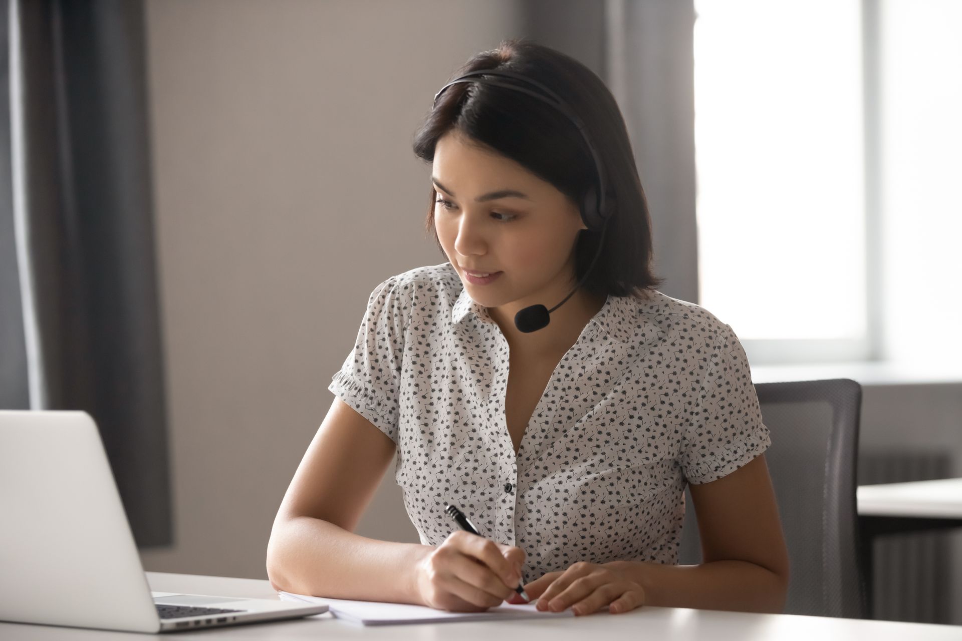 A person wearing a headset works at a desk, looking at a laptop while writing on a paper.