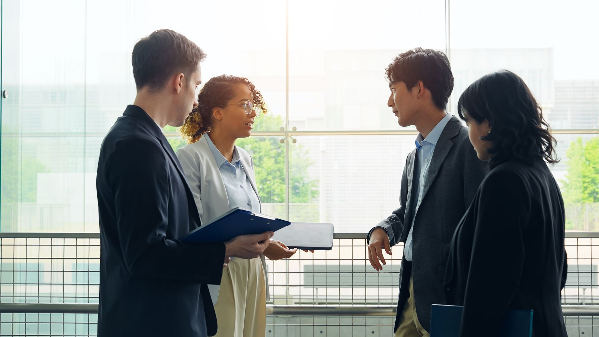 Four business professionals in formal attire confer while standing in a bright, modern office hallway.