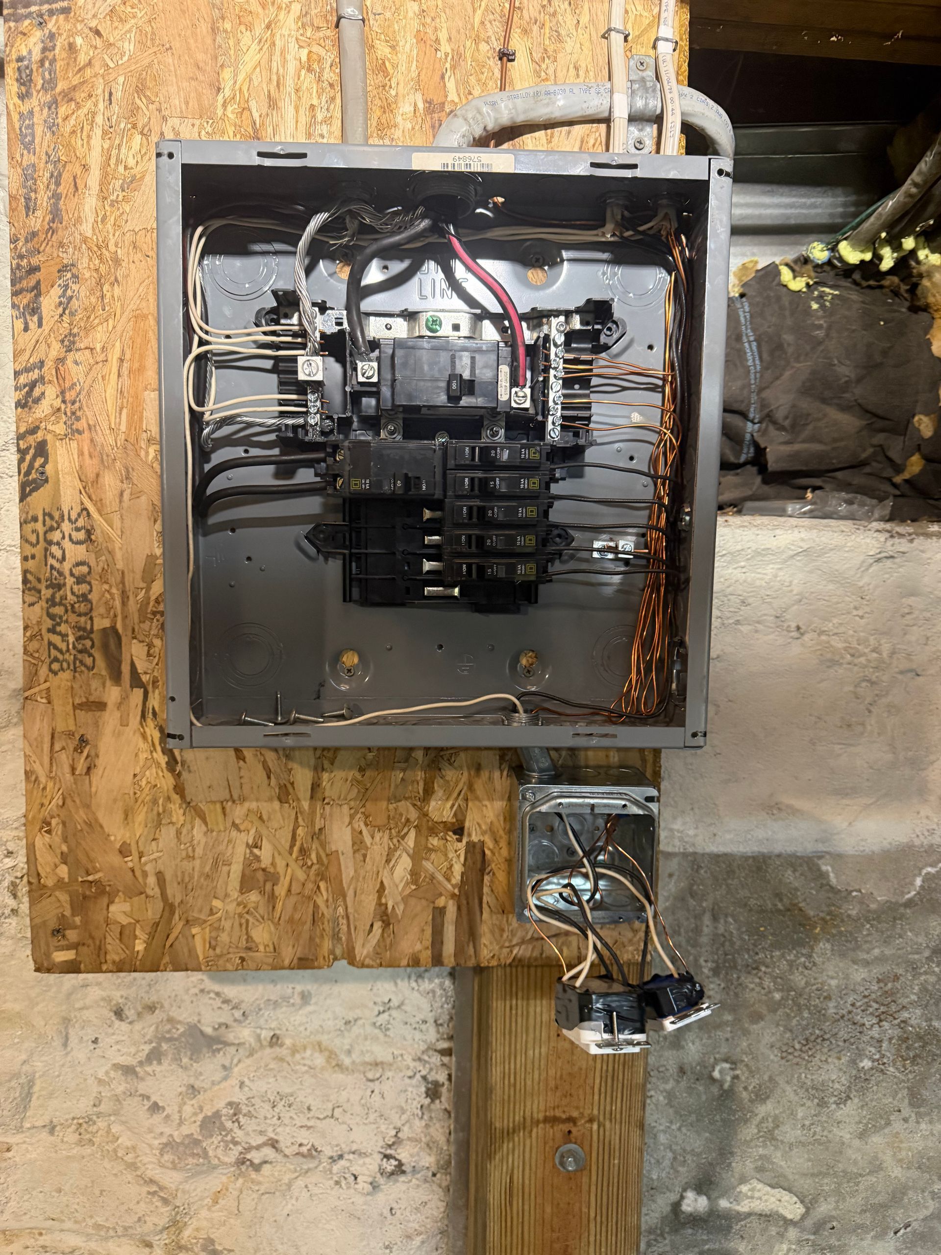 An open electrical panel mounted on a wooden board, with wires and two hanging outlets below.