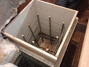 A square wooden formwork box containing reinforced steel bars, set into a dirt foundation for concrete pouring.