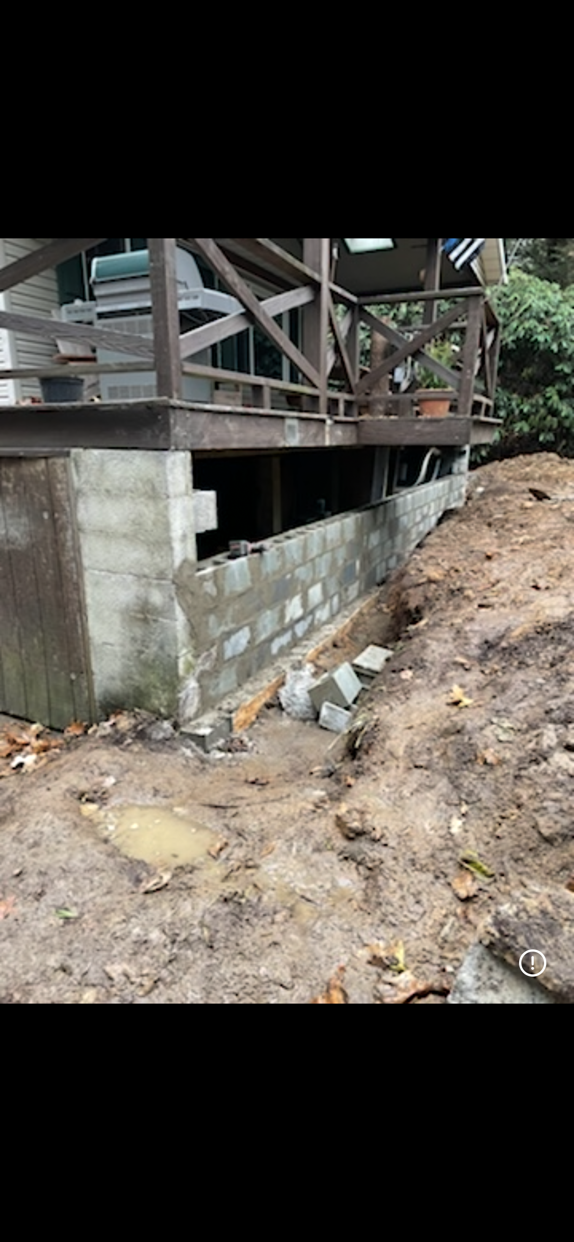 Concrete blocks being installed to build or repair a retaining wall beneath a wooden deck on a sloping, muddy lot.