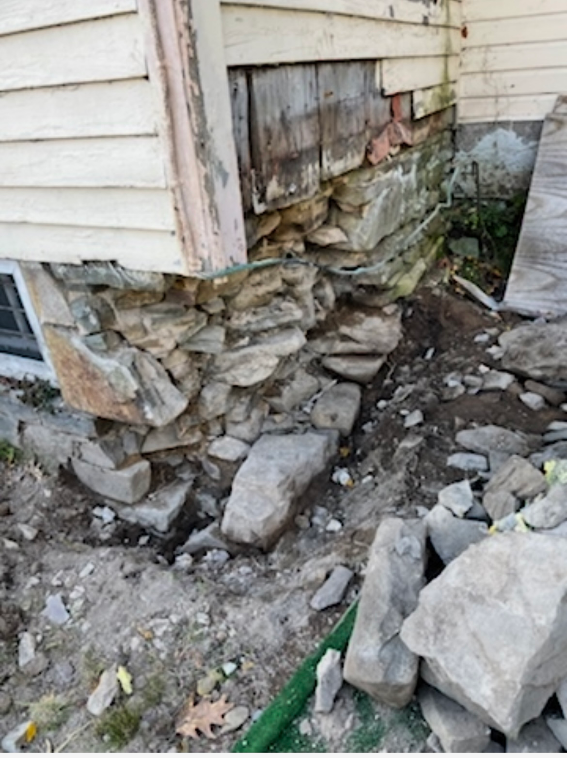 The stone foundation corner of a house is crumbling, with loose rocks and dirt on the ground below.